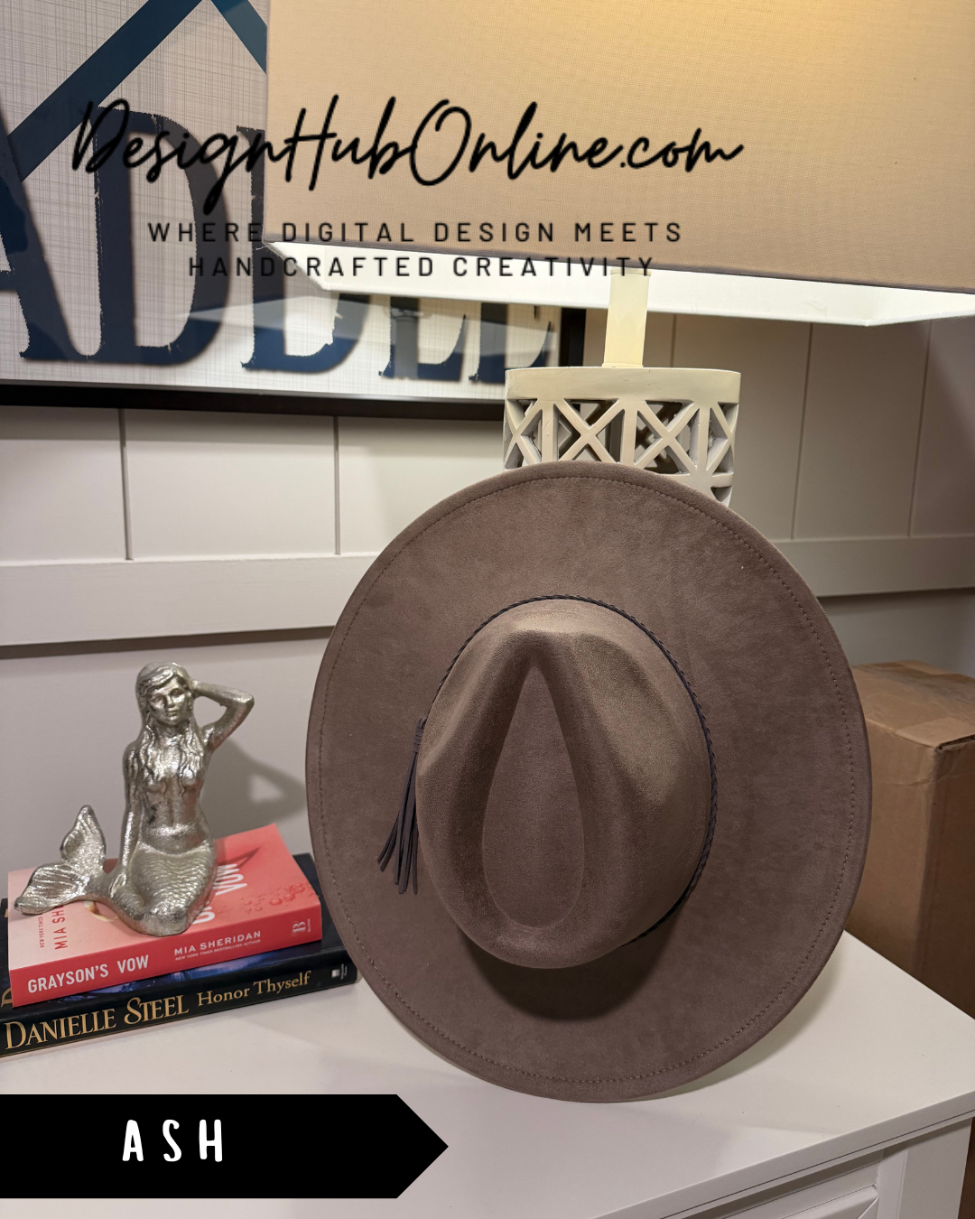 a brown fedora hat placed on a white surface, with a small statue of a mermaid next to it. There is also a book titled "Diana Steel" and a lamp in the background.