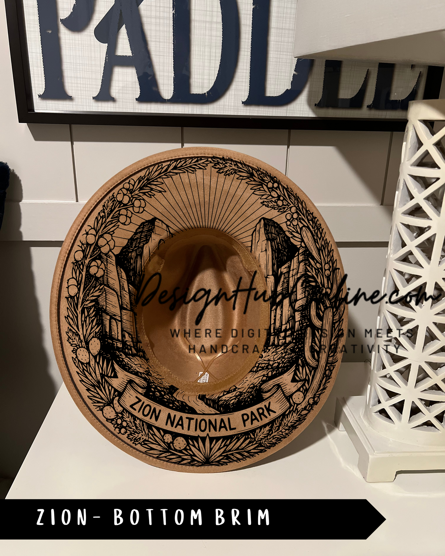 A brown hat with a decorative design, labeled "ZION NATIONAL PARK" and "ZION-BOTTOM BRIM", is displayed on a shelf.