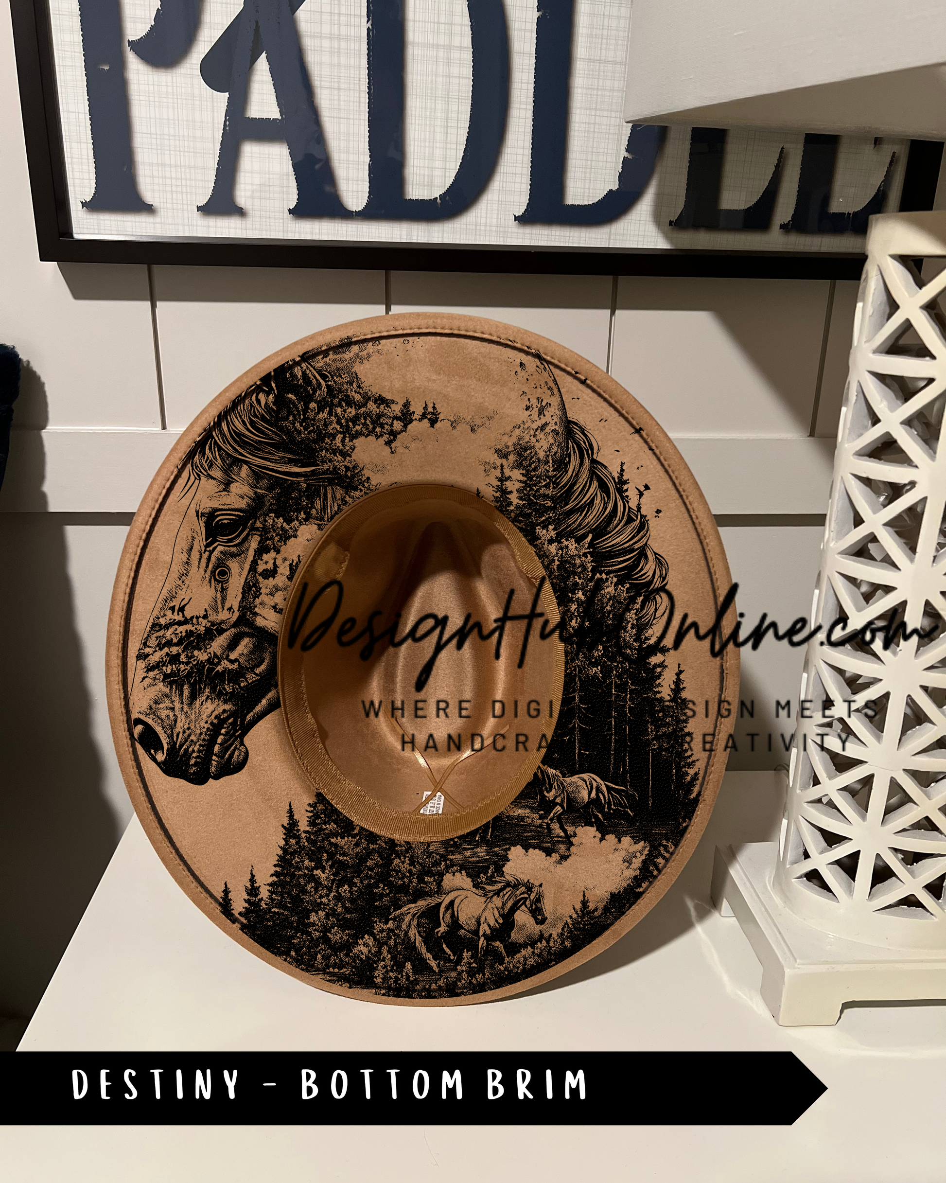 A brown cowboy hat with a black and white illustration of a horse and forest on the front, displayed on a shelf.