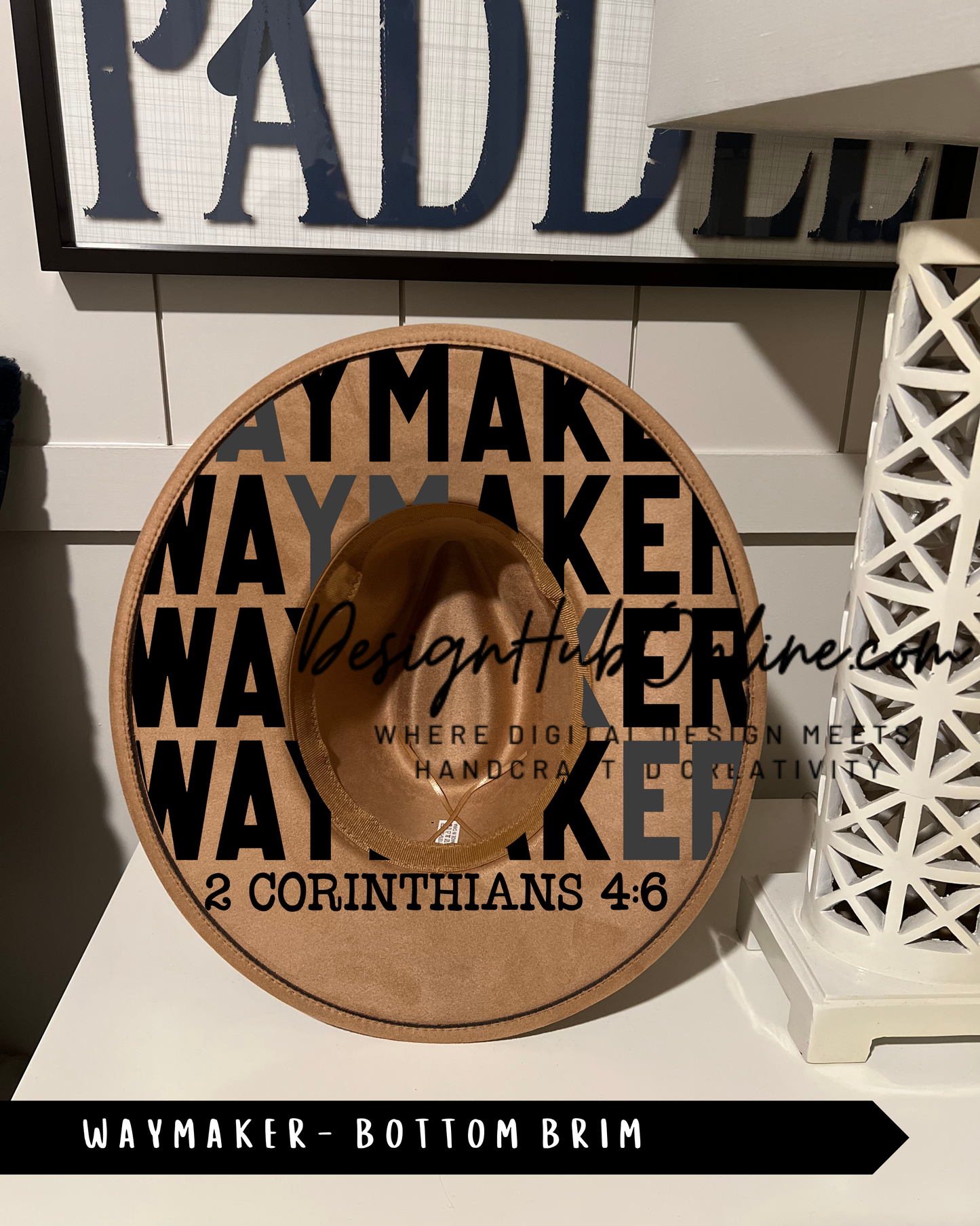 a brown Waymaker hat with the text "WAYMAKER" and "2 CORINTHIANS 4:6" printed on it. The hat is placed on a white surface, and there is a sign with the text "PADDLER" in the background.