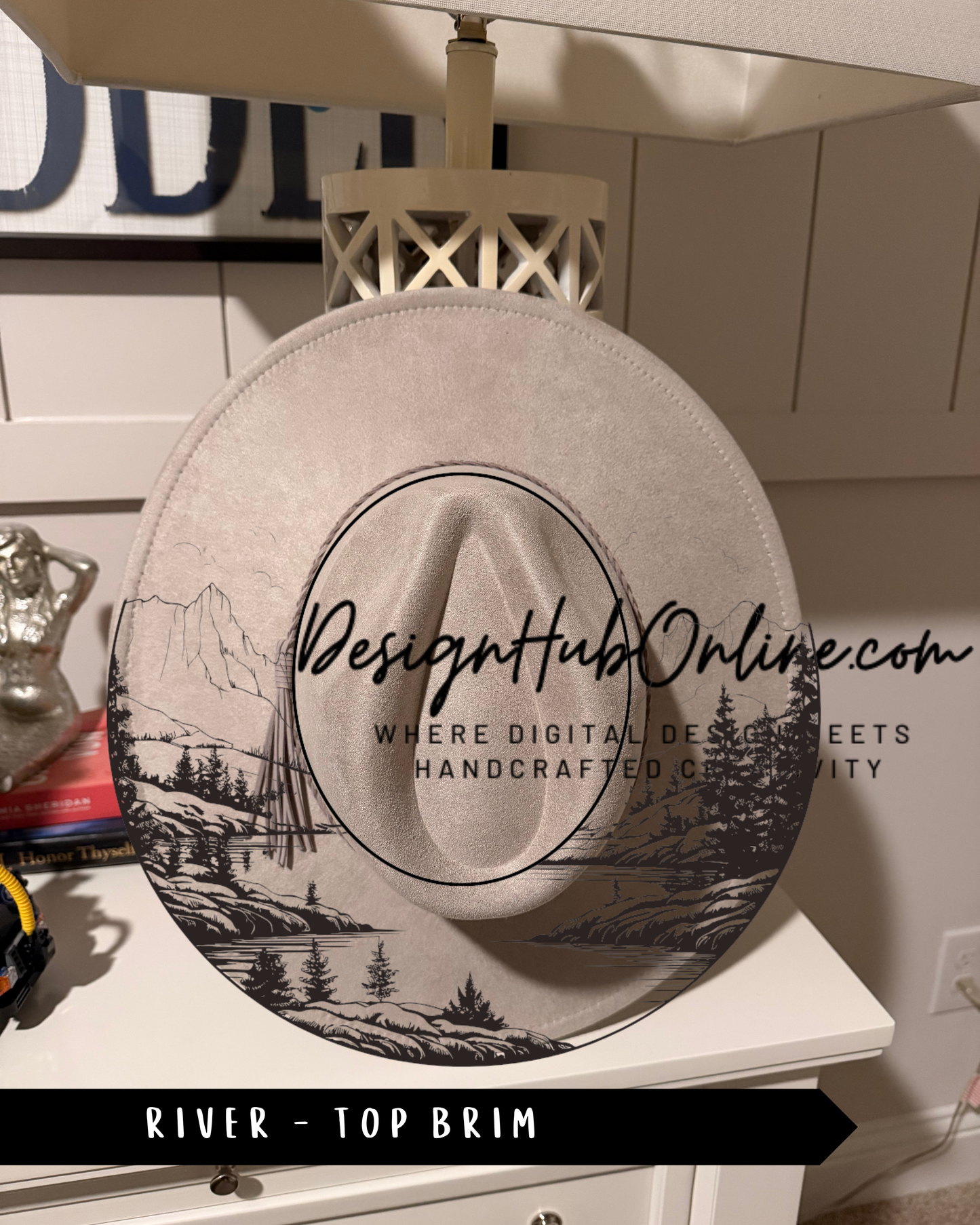 A white hat with a black and white illustration of a river and mountains on the front, placed on a white dresser.