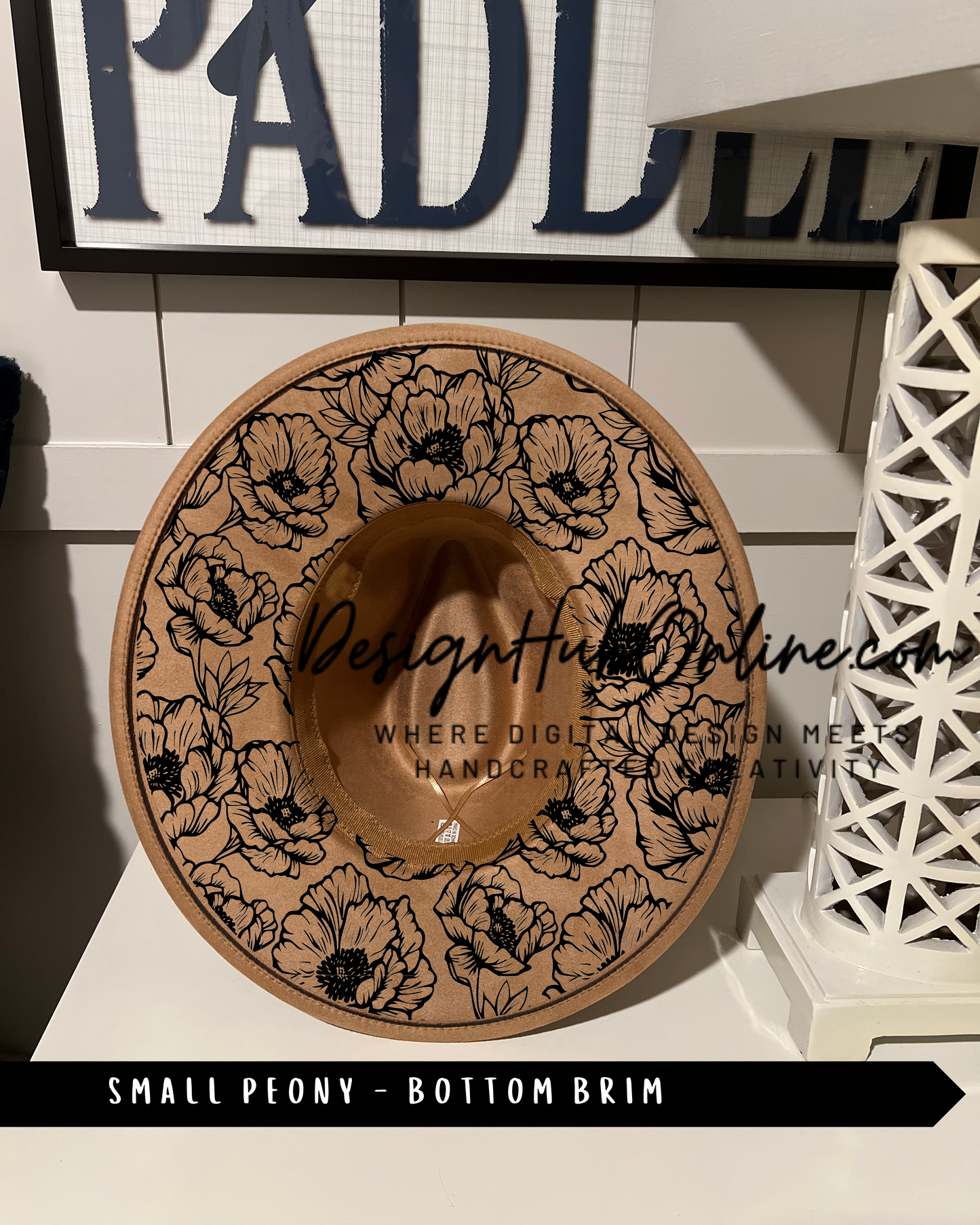 A small peony-patterned hat with a bottom brim design, displayed on a shelf against a white wall with a sign that reads 'PADLL'.