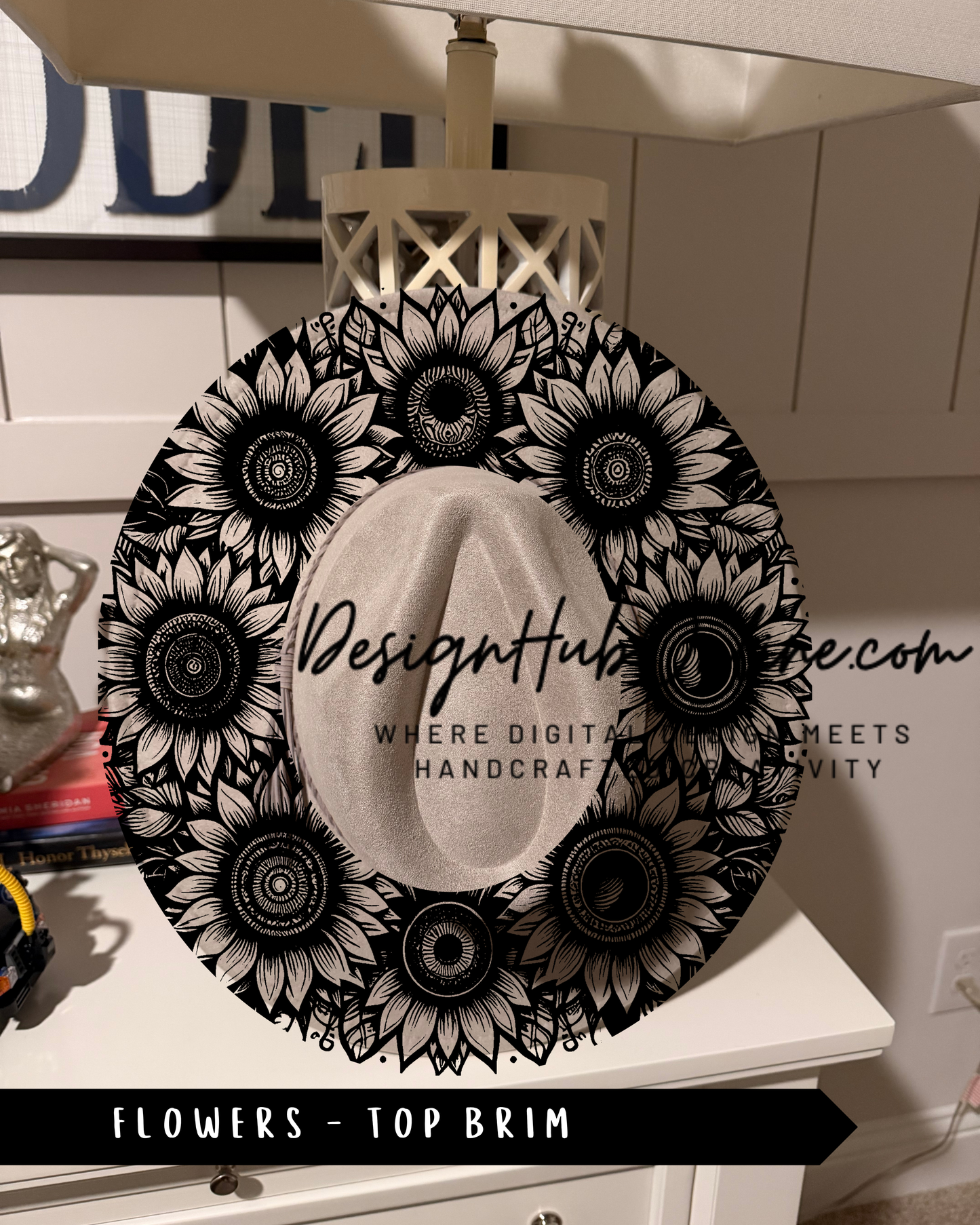 A decorative hat with a floral pattern, featuring a large sunflower design surrounded by smaller sunflower designs. The hat is placed on a white dresser, and there is a sign with text that reads "Design Hub" and "Where digital meets handmade".