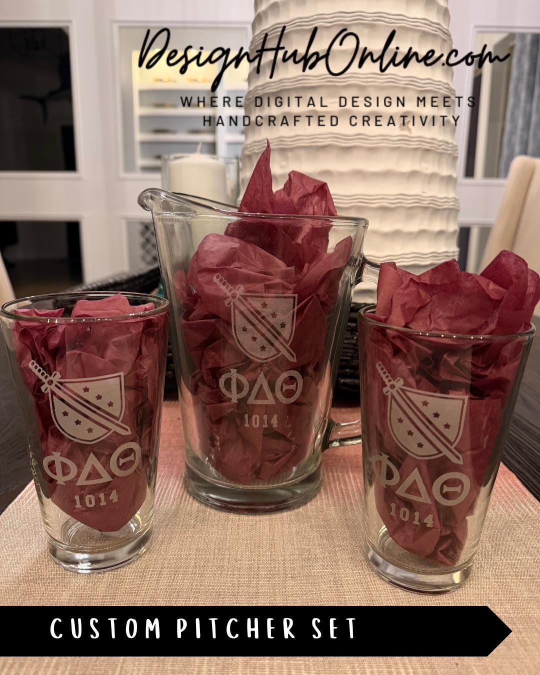 A set of three glass pitchers with the Greek letters ΦΦΦ displayed on them, along with a decorative red tissue paper. The pitchers are arranged on a table, and there is a sign in the background that reads "DesignHUBOnline.com".