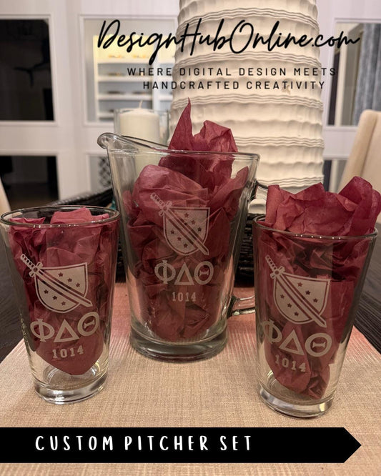 A set of three glass pitchers with the Greek letters ΦΦΦ displayed on them, along with a decorative red tissue paper. The pitchers are arranged on a table, and there is a sign in the background that reads "DesignHUBOnline.com".