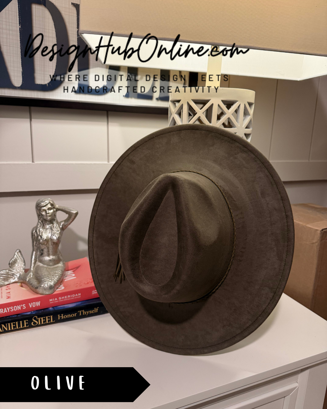 a brown fedora hat placed on a white surface, with a small silver figurine of a mermaid to the left. There is also a book titled "Mia Sheeran Honor Thyself" on the surface.