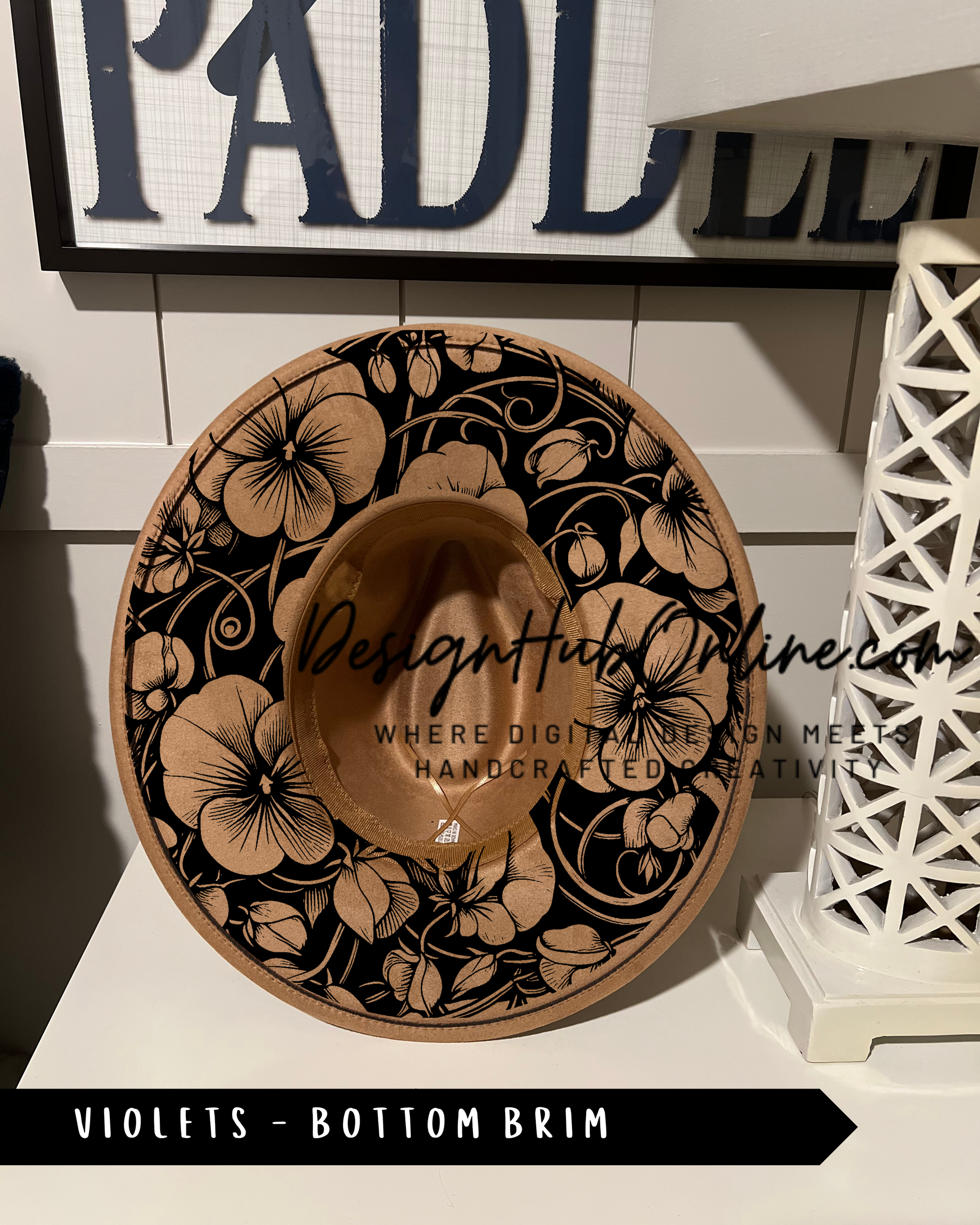 A decorative hat with a floral pattern, featuring a mix of black and gold colors. The hat is placed on a white surface, with a sign in the background that reads "PADLLES".