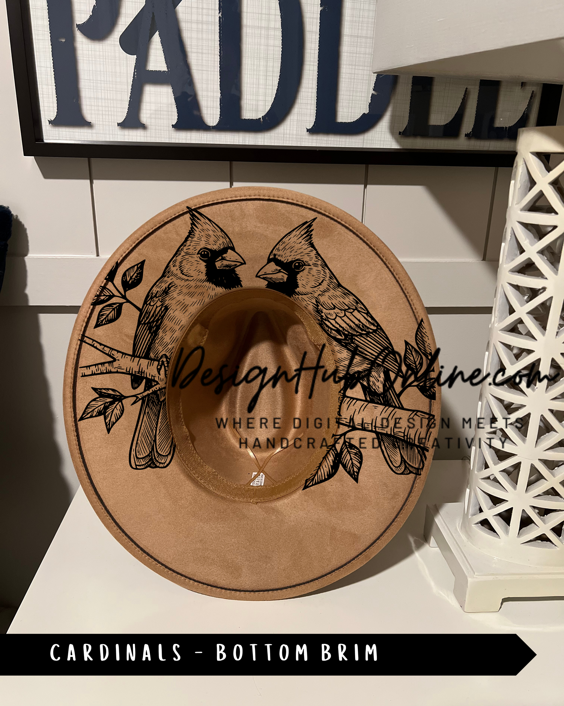 A brown cowboy hat with a design of two black birds, likely cardinals, on the front. The hat is placed on a white surface, with a sign that reads "PADLL" in the background.